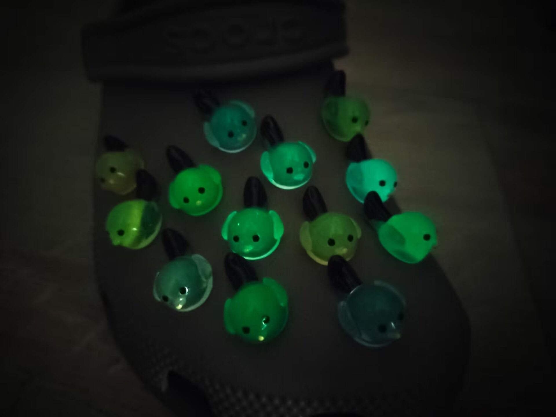 Taupe-colored shoe shoe adorned with glow-in-the-dark bird charms. Each charm features a small, round bird with a beak and black wings, glowing softly in various colors. The shoe is positioned outdoors against a backdrop of green foliage.