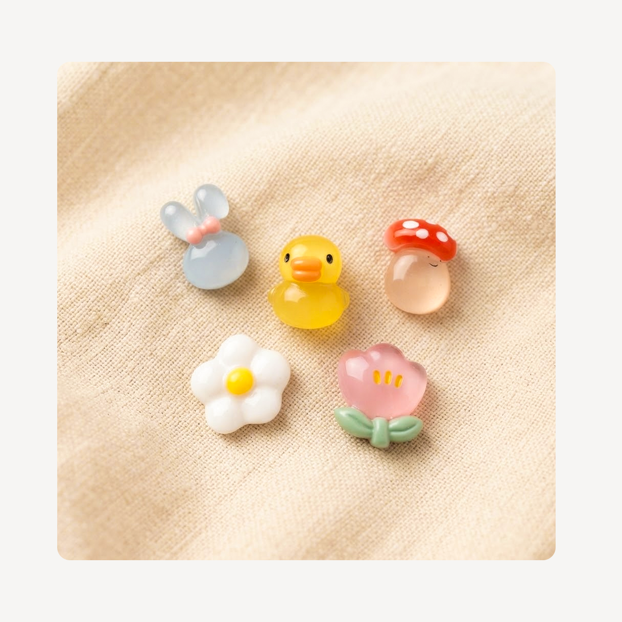 Five small decorative clog charms arranged on a beige fabric background: a pale blue bunny, a yellow duck, a red-and-white mushroom, a white flower with a yellow center, and a pink flower with green leaves.