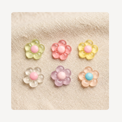 Six small translucent flower-shaped clog charms arranged in two rows on a beige fabric background. The flowers are in pastel shades including yellow-green, pink, pale yellow, clear with pink center, lavender, peach, and one with a light blue center.