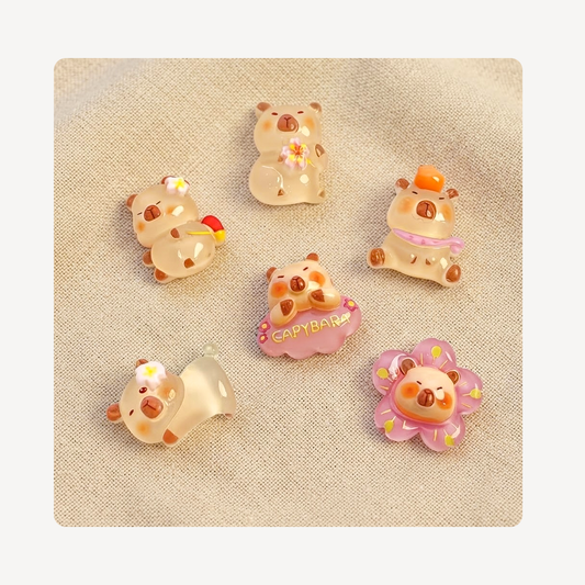Six small decorative animal-shaped clog charms arranged on a beige fabric background. The characters are cream and peach with pastel accents including pink, yellow, orange, and purple.