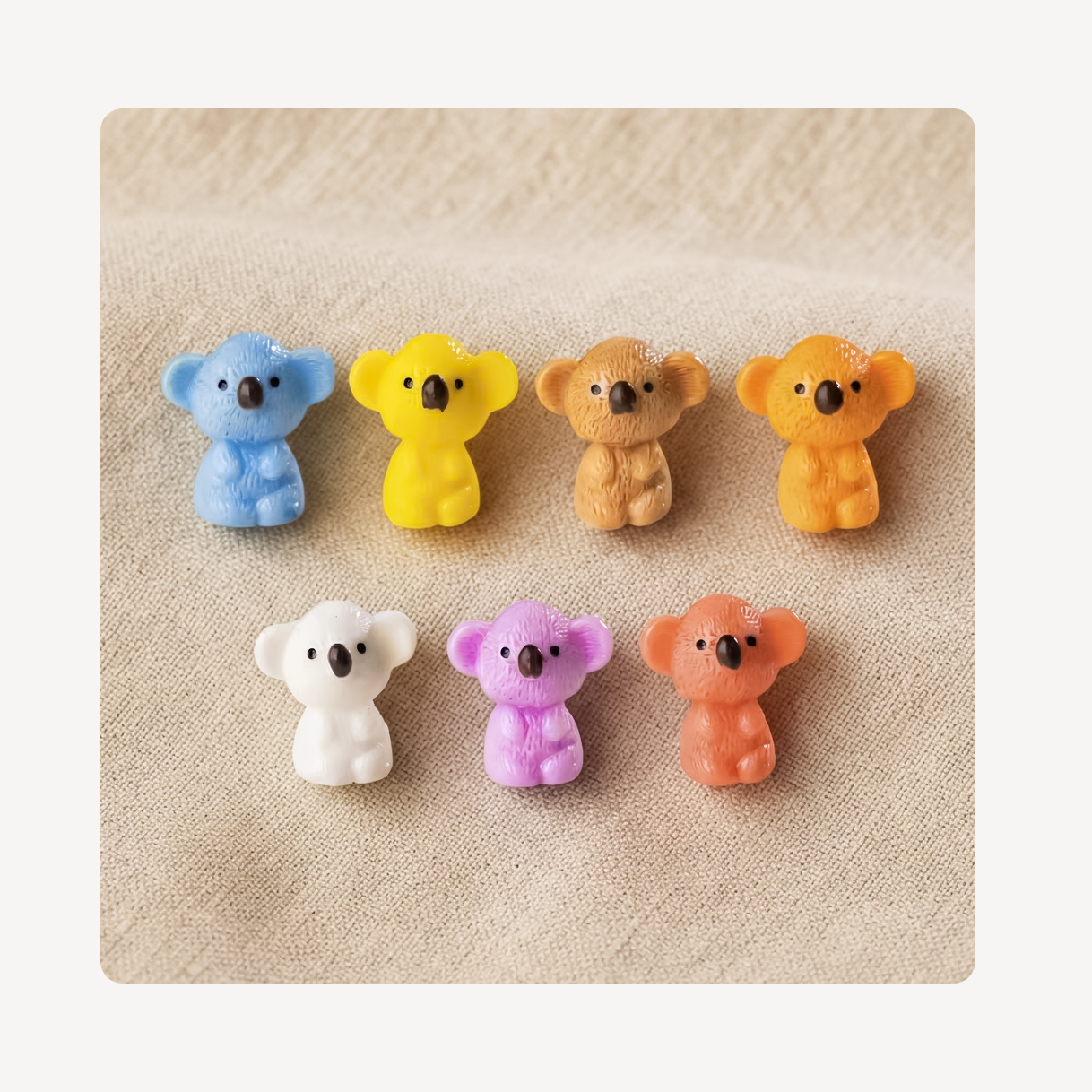 Six small koala-shaped clog charms in assorted colors, including blue, yellow, tan, orange, white, purple, and peach, arranged in two rows on a textured beige fabric background.