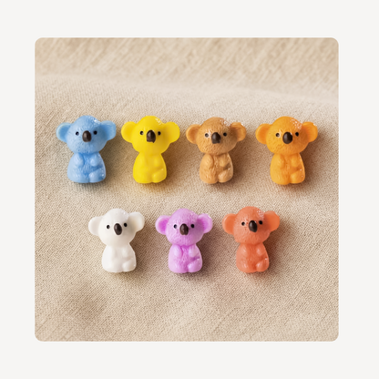 Six small koala-shaped clog charms in assorted colors, including blue, yellow, tan, orange, white, purple, and peach, arranged in two rows on a textured beige fabric background.