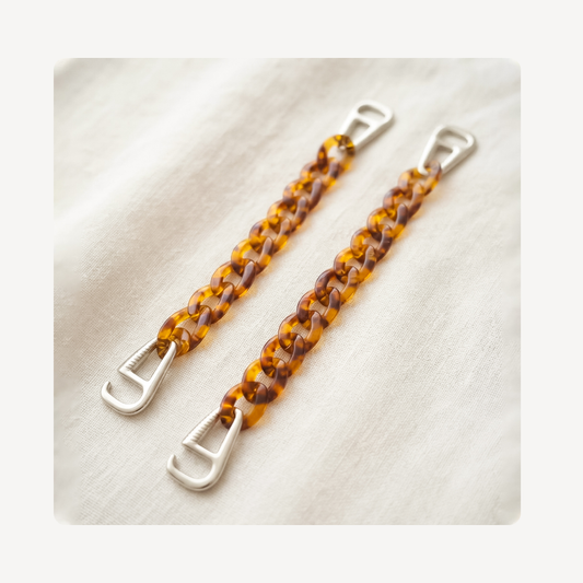 Two amber and brown translucent chain link clog chains with silver metal lobster clamps, displayed on a soft white textured fabric background.