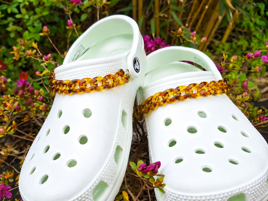 A pair of white Croc-style clogs with chunky amber-brown chain straps, photographed outdoors among green foliage and small pink flowers.