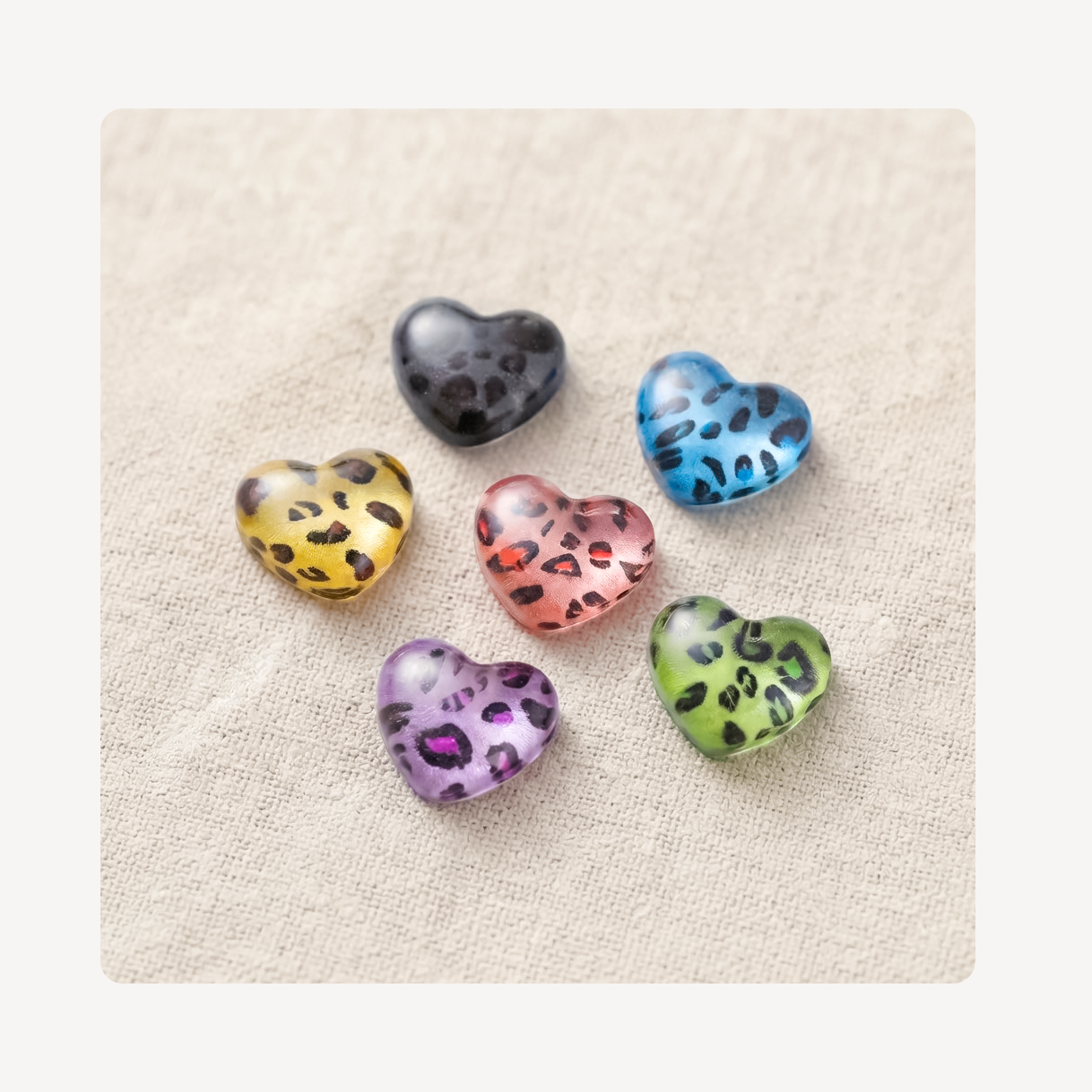 Five small heart-shaped clog charms with black leopard spots in yellow, blue, pink, green, and purple, arranged on a beige textured fabric background.