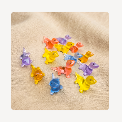 A group of small translucent lizard-shaped clog charms in assorted colors including yellow, blue, purple, orange, and pink, arranged on a beige textured fabric background.