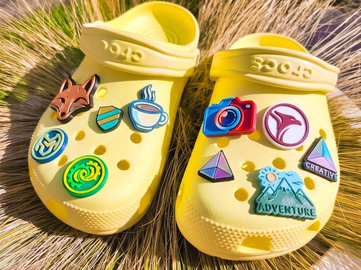 A pair of yellow Crocs clogs decorated with colorful, custom logo charms, resting on a bed of dry grass. The charms include a fox head, a coffee cup, a mountain with a sun and the word 'ADVENTURE', a camera, and geometric shapes.