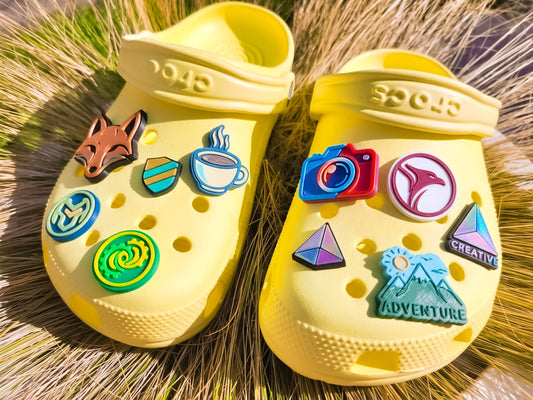 A pair of yellow Crocs clogs decorated with colorful, custom logo charms, resting on a bed of dry grass. The charms include a fox head, a coffee cup, a mountain with a sun and the word 'ADVENTURE', a camera, and geometric shapes.
