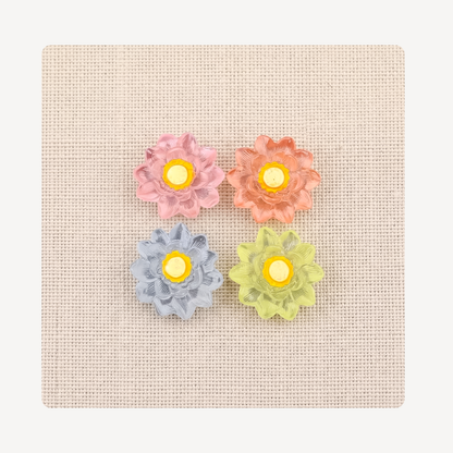 Four flower-shaped clog charms in pastel pink, peach, light blue, and lime green, arranged on a square beige textured fabric background.