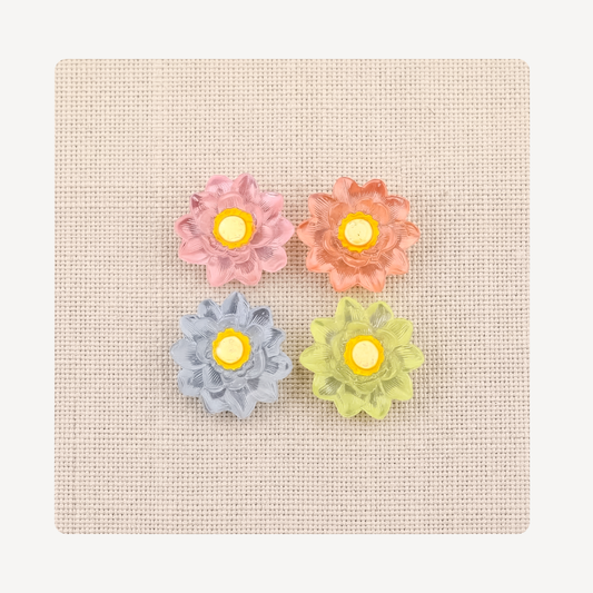 Four flower-shaped clog charms in pastel pink, peach, light blue, and lime green, arranged on a square beige textured fabric background.