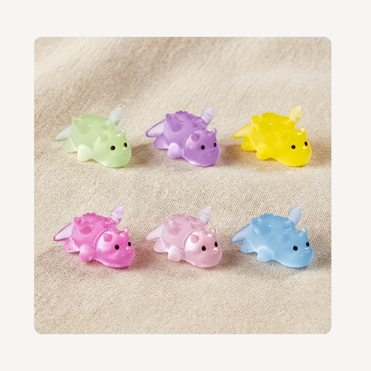 Six small translucent dragon clog charms in pastel colors—green, purple, yellow, pink, light pink, and blue—arranged on a beige textured fabric background.