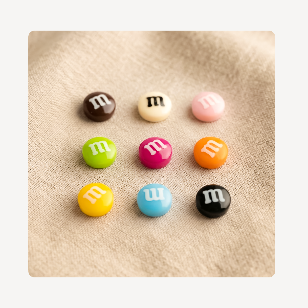 Nine small round candy-like clog charms with white lowercase 'm' logos arranged in a 3-by-3 grid on a beige textured fabric background. The charms come in brown, cream, pink, green, magenta, orange, yellow, light blue, and black.