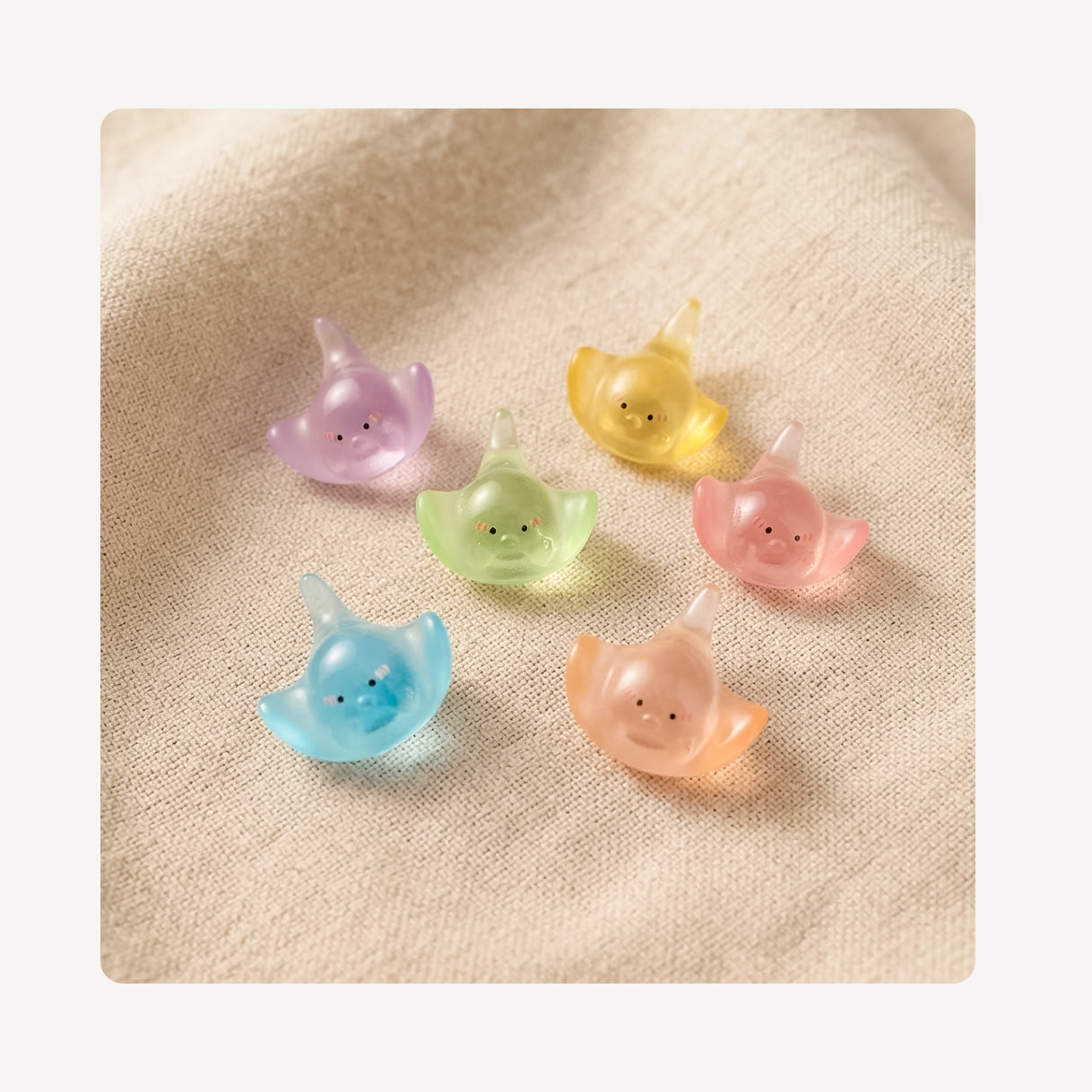 Six small translucent manta ray clog charms in pastel colors (purple, yellow, green, pink, and blue) arranged on a textured beige fabric background.