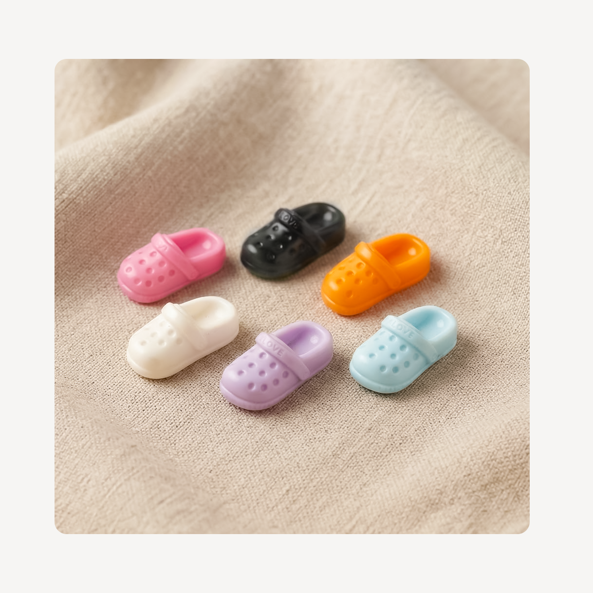 Five miniature clog-style clog charms in pink, black, orange, cream, lavender, and light blue, arranged on a beige textured fabric background.