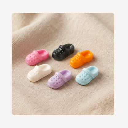 Five miniature clog-style clog charms in pink, black, orange, cream, lavender, and light blue, arranged on a beige textured fabric background.