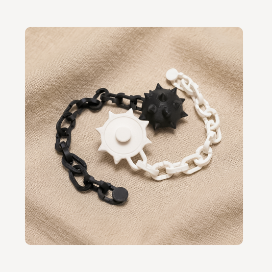 A clog chain charm set featuring a black metal-link style chain with a black spiked ball and a white chain with a white spiked ball, arranged on a beige textured fabric background.