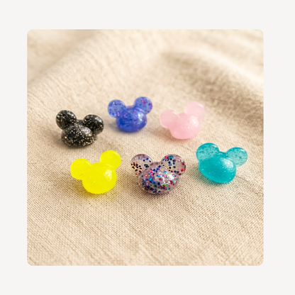 Six small Mickey Mouse-shaped clog charms with different colors and glitter finishes, arranged on a beige textured fabric background.
