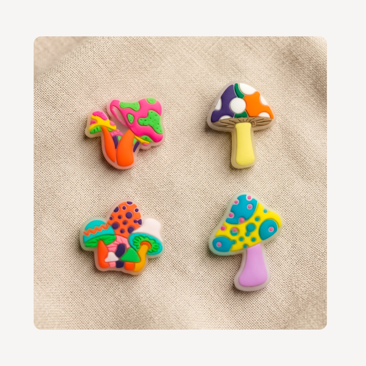 Four colorful mushroom-shaped clog charms arranged on a beige textured fabric background.