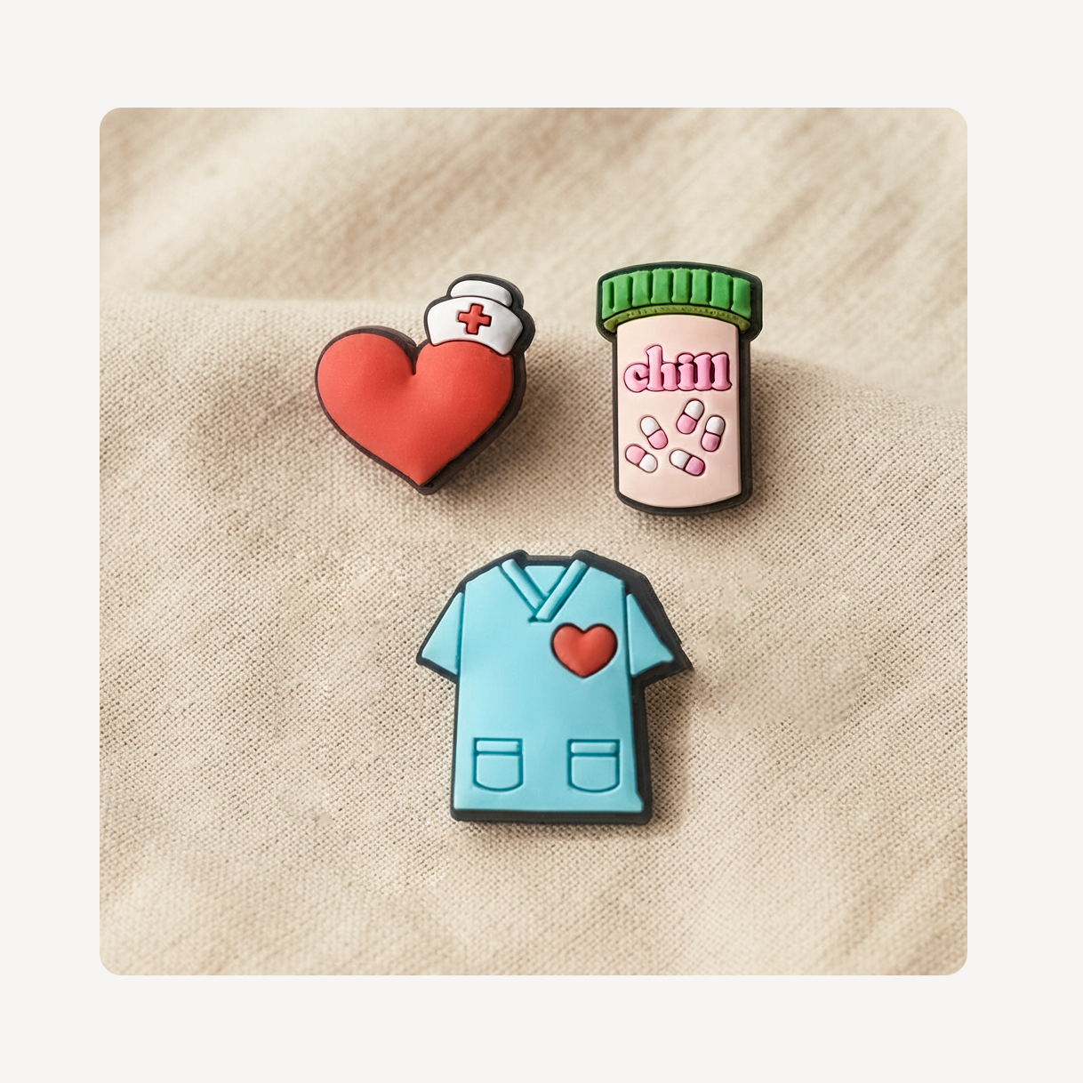 Three medical-themed clog charms shaped like a red heart with a nurse cap, a pill bottle, and a light blue scrub top with a red heart, displayed on a beige textured fabric background.