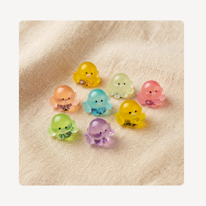 Eight small translucent octopus-shaped clog charms in assorted pastel colors, including yellow, pink, blue, green, and purple, arranged on a beige textured fabric background.