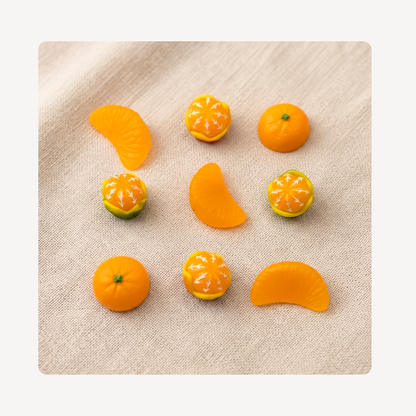 Nine miniature orange-themed clog charms arranged on a beige linen-textured surface. The set includes whole oranges, orange slices, and oranges with white starburst patterns resembling citrus pith or decorative detailing.