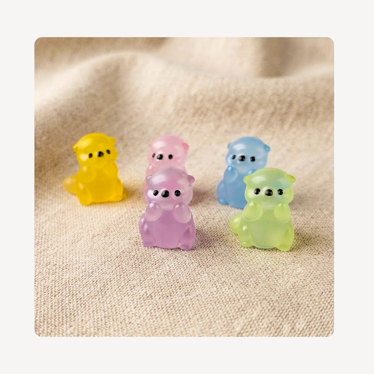 Five small translucent otter-shaped clog charms in yellow, pink, blue, and green, arranged on a beige textured fabric background.