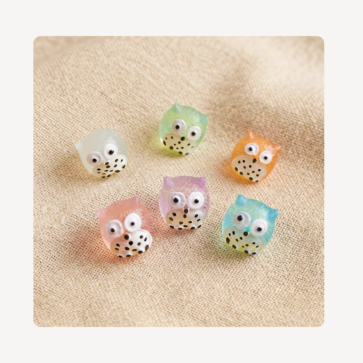 Six small owl-shaped clog charms in pastel colors including white, mint green, orange, pink, lavender, and light blue, arranged on a beige textured fabric background.