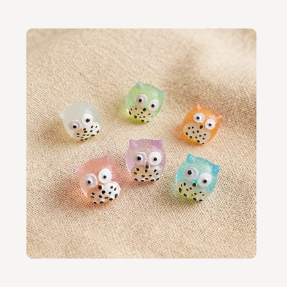 Six small owl-shaped clog charms in pastel colors including white, mint green, orange, pink, lavender, and light blue, arranged on a beige textured fabric background.