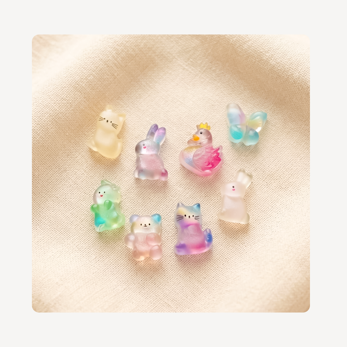 Seven small translucent clog charms shaped like animals, including bears, a rabbit, a duck, a butterfly, and cats, arranged on a textured beige fabric background.