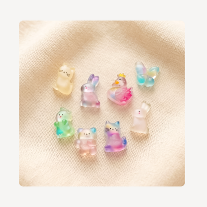 Seven small translucent clog charms shaped like animals, including bears, a rabbit, a duck, a butterfly, and cats, arranged on a textured beige fabric background.