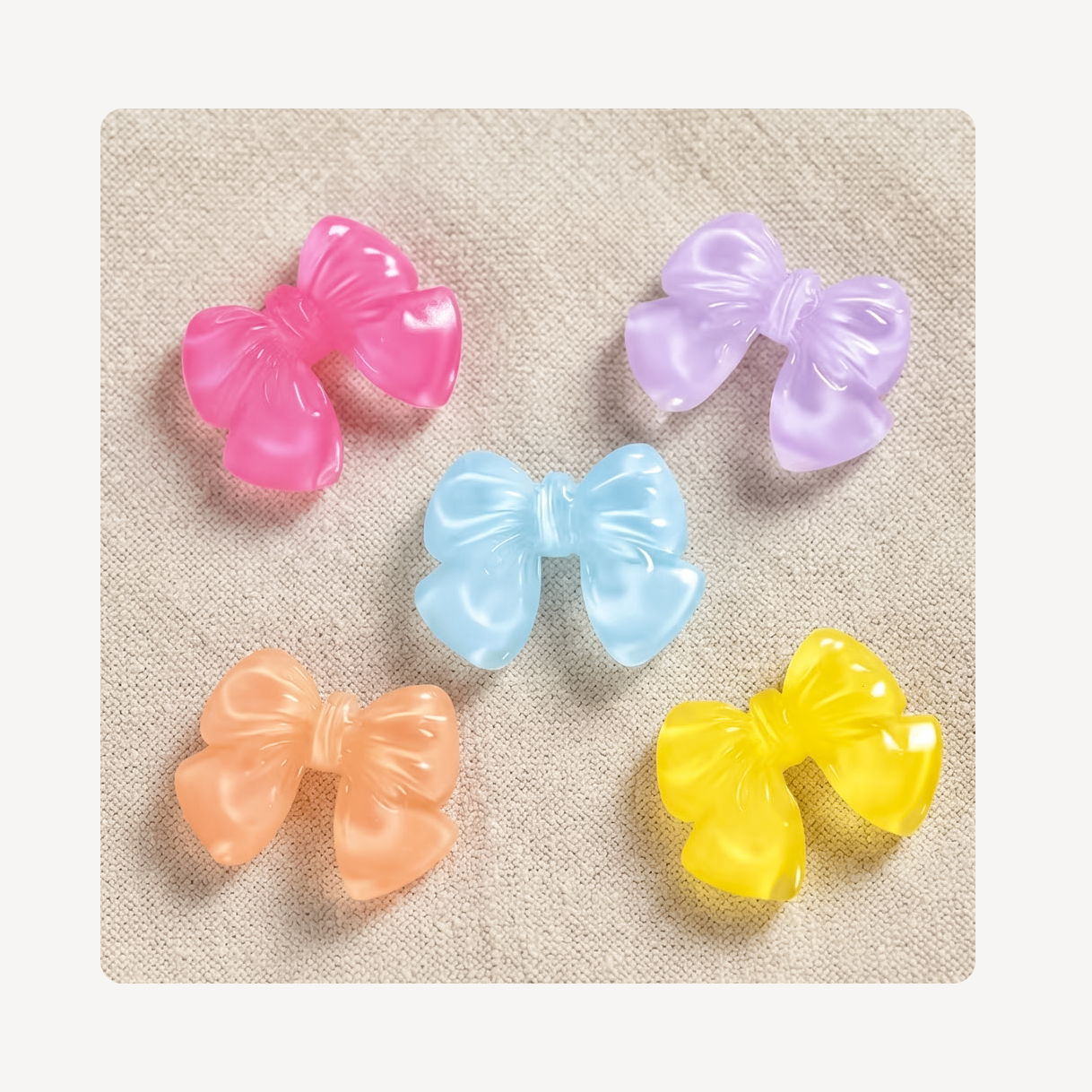Five glossy translucent bow clog charms in pink, purple, light blue, peach, and yellow, arranged on a textured beige fabric background.