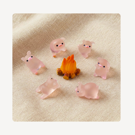 Six small translucent pink piggy clog charms arranged around a tiny orange campfire on a beige fabric background.
