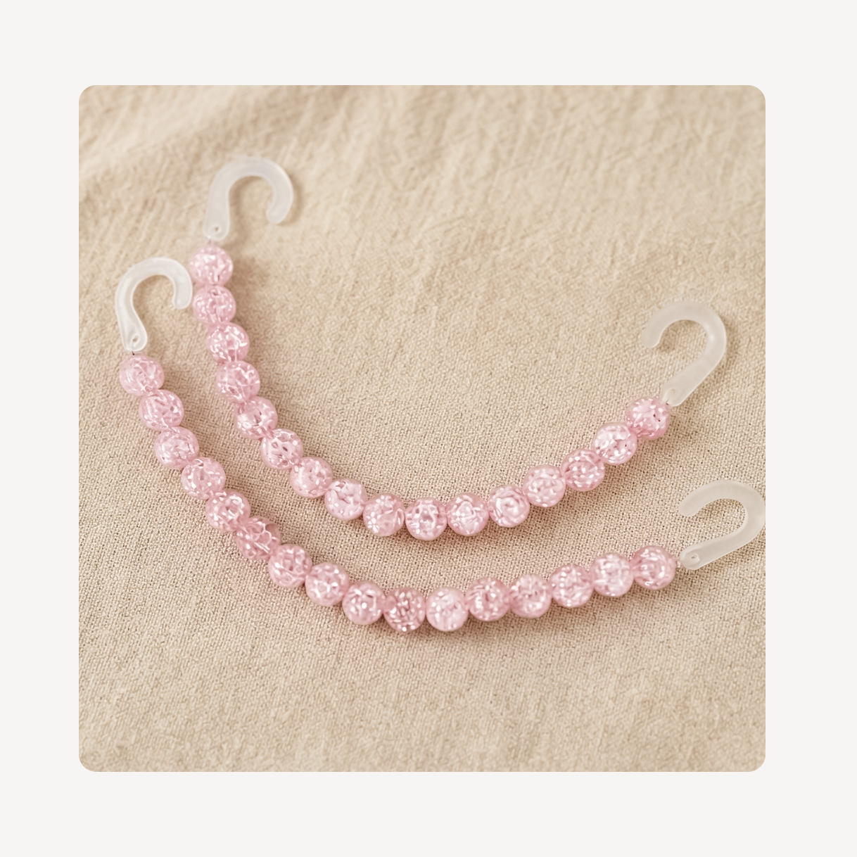 Two pink beaded clog chains with clear plastic hooks, arranged on a textured beige background.
