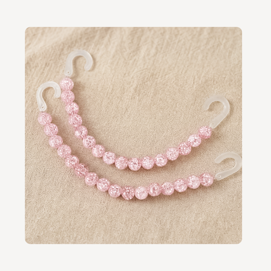 Two pink beaded clog chains with clear plastic hooks, arranged on a textured beige background.