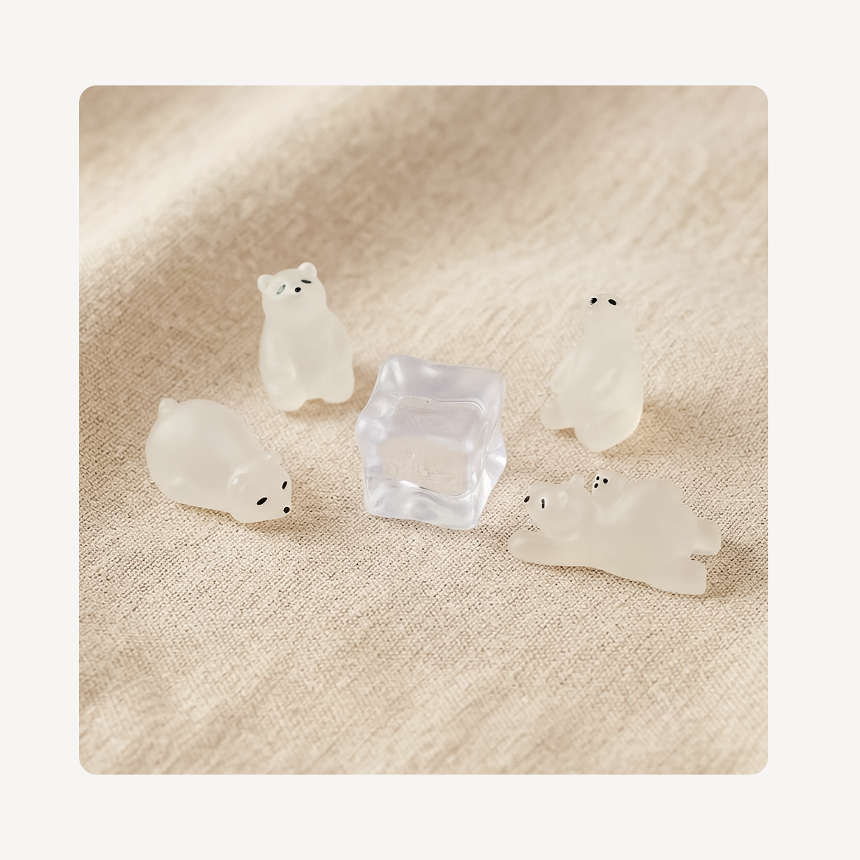 Four small translucent white bear-shaped clog charms arranged around a clear square ice cube on a beige textured fabric background.