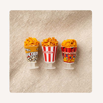 Three miniature popcorn bucket clog charms filled with yellow-orange popcorn, displayed on a textured beige fabric background.