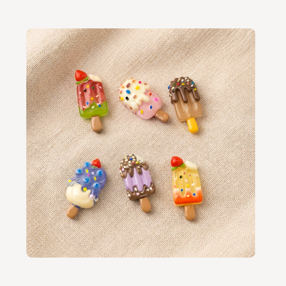 Six miniature popsicle-shaped clog charms arranged on a textured beige fabric background.