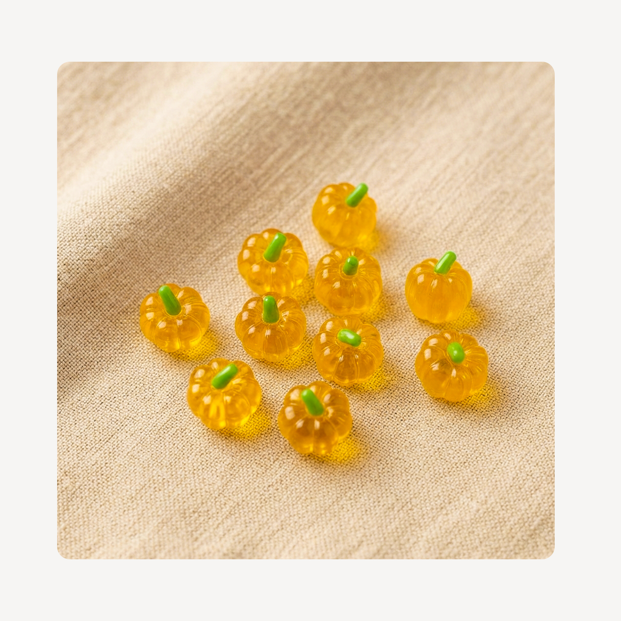 Nine small translucent orange pumpkin-shaped clog charms with green stems, arranged on a beige textured fabric background.