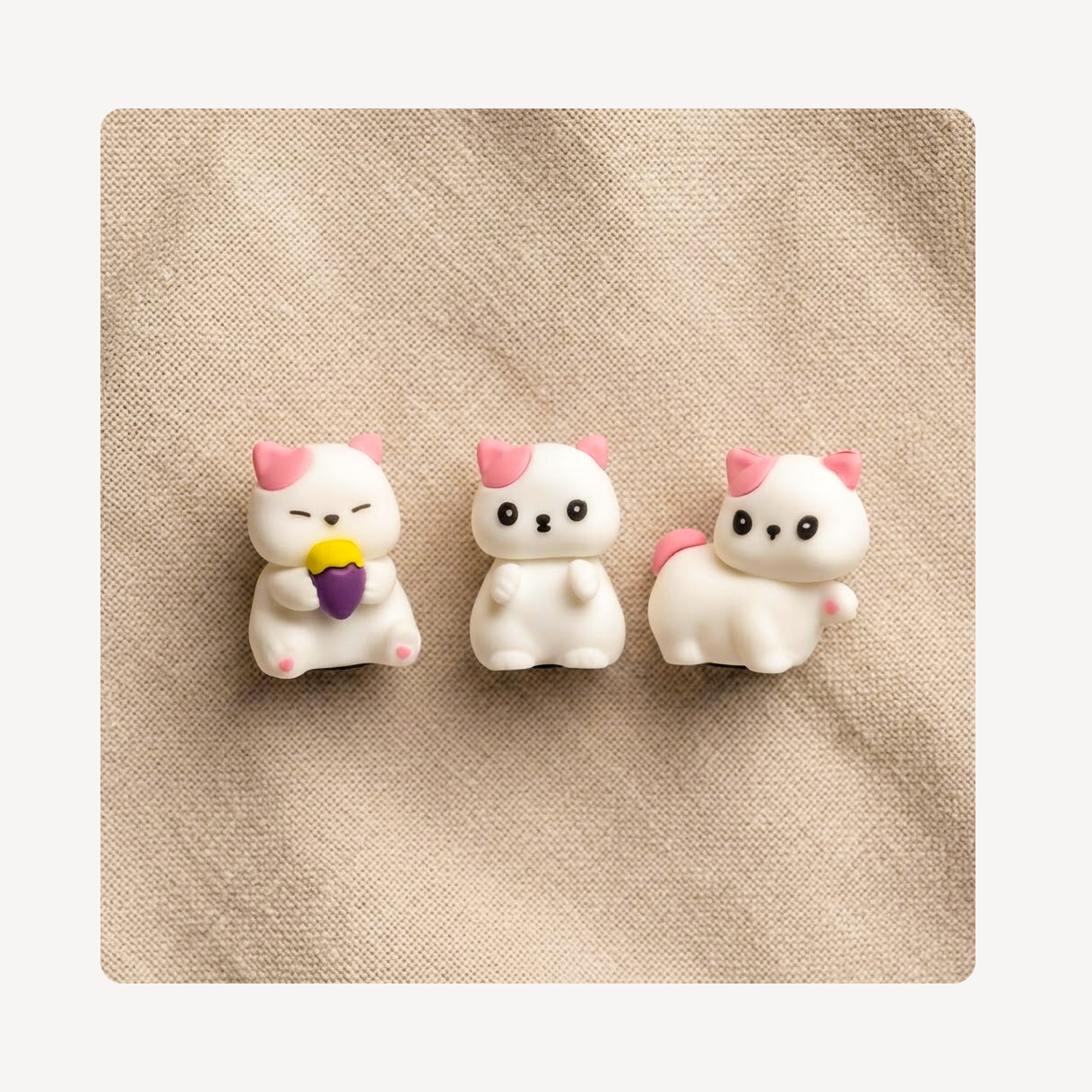 Three small white cat clog charms with pink ears and tails, arranged side by side on a beige textured fabric background.