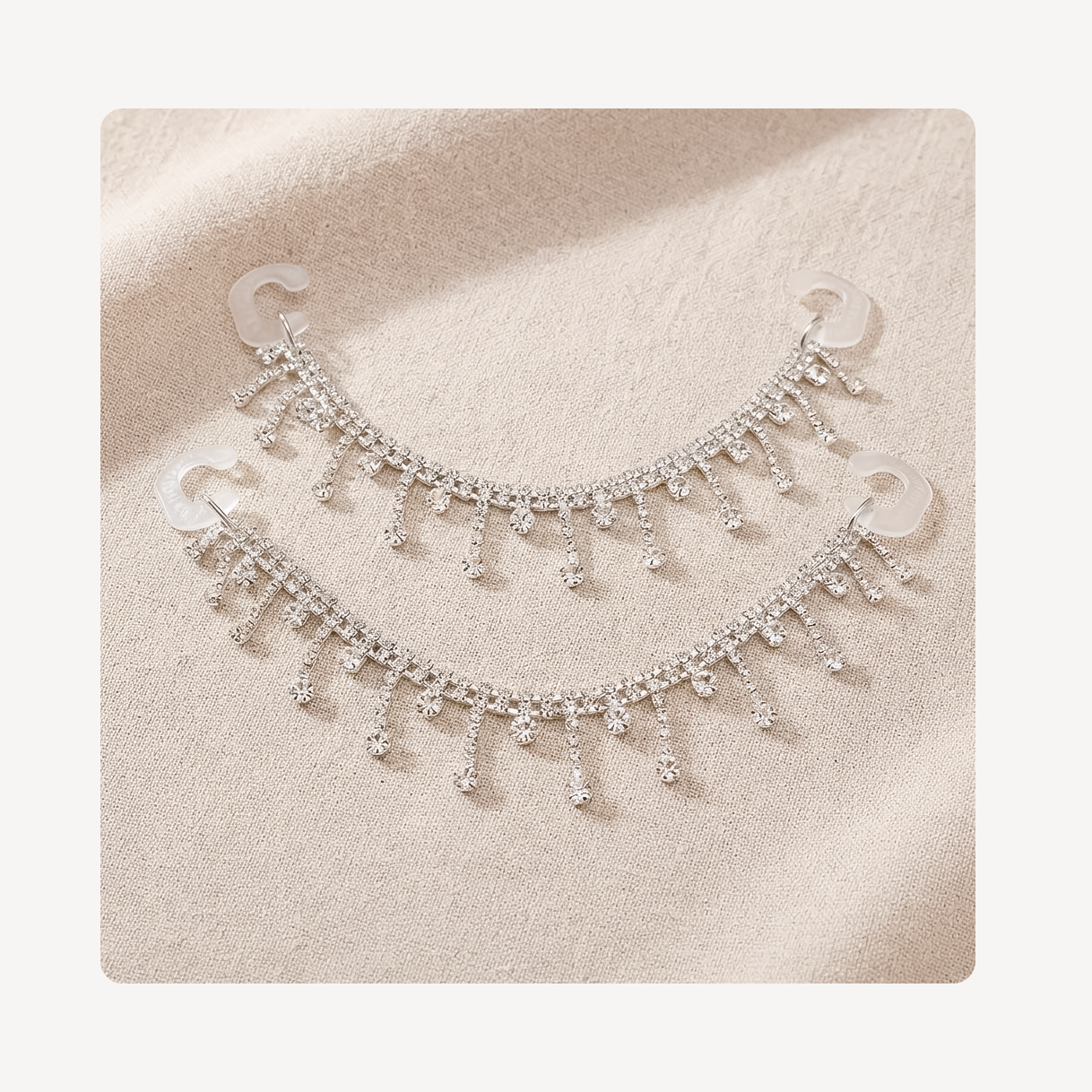 A pair of silver-toned crescent-shaped clog chains adorned with dangling rhinestone fringe, displayed on a textured beige fabric background.