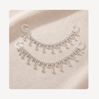 A pair of silver-toned crescent-shaped clog chains adorned with dangling rhinestone fringe, displayed on a textured beige fabric background.