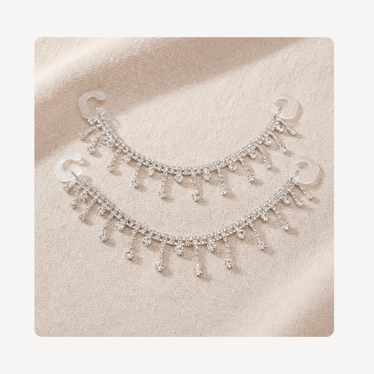 A pair of silver-toned crescent-shaped clog chains adorned with dangling rhinestone fringe, displayed on a textured beige fabric background.