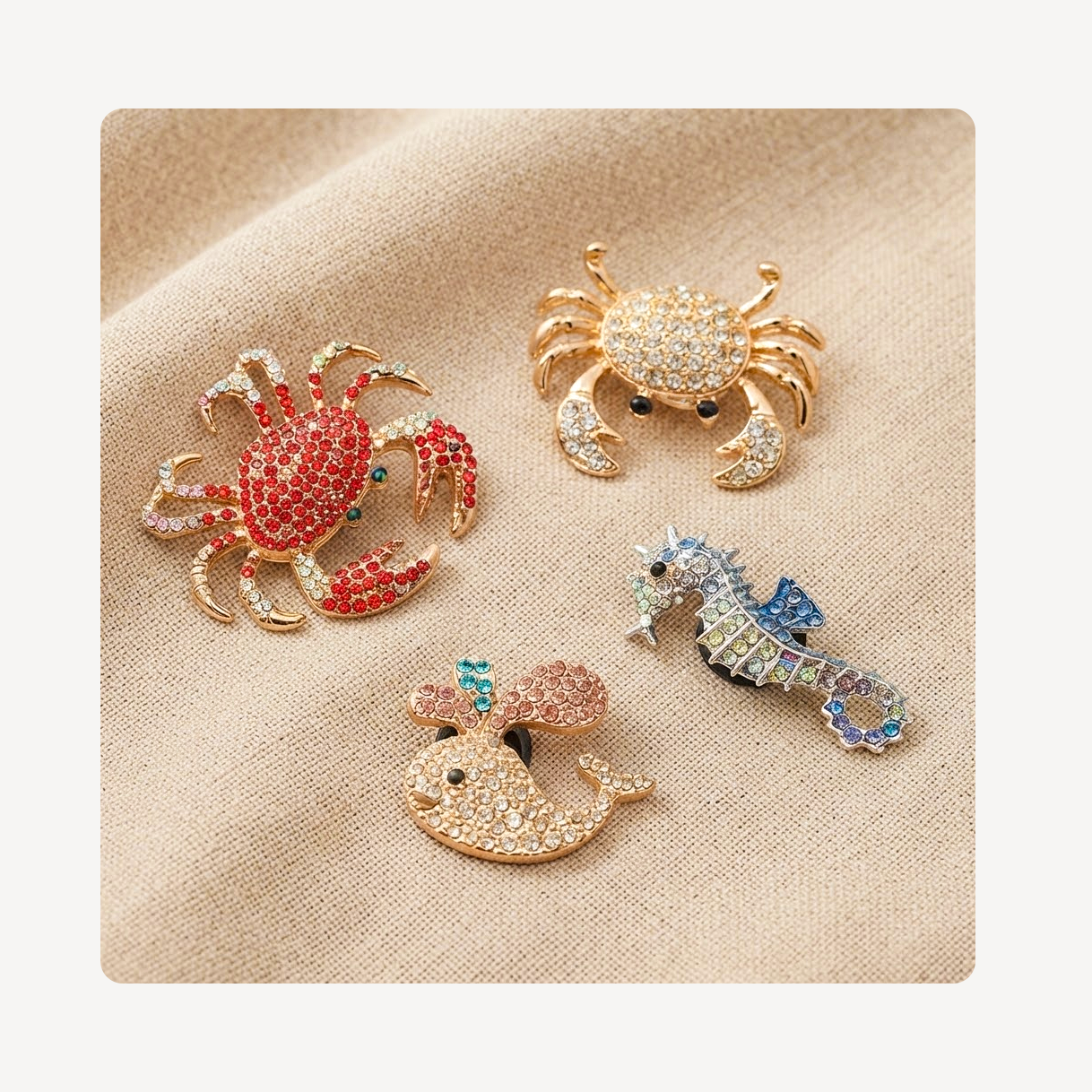 Four nautical-themed rhinestone clog charms on a beige textured fabric background: a red and gold crab, a gold crab with clear stones, a blue and silver seahorse, and a gold whale with pink and blue accents.