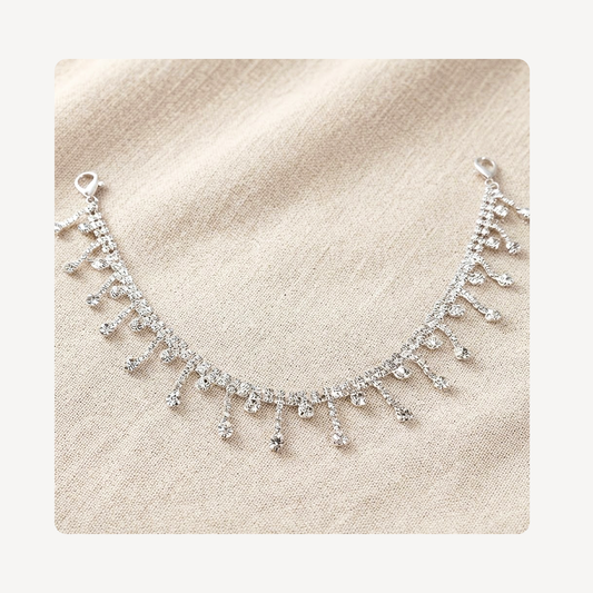 A silver-toned crystal fringe shoe chain displayed on a textured beige fabric background.