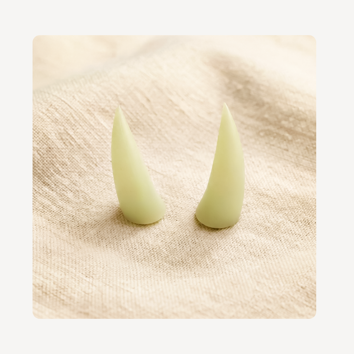 A pair of pale yellow-green pointed rhino horn clog charms standing upright on a textured beige fabric surface.