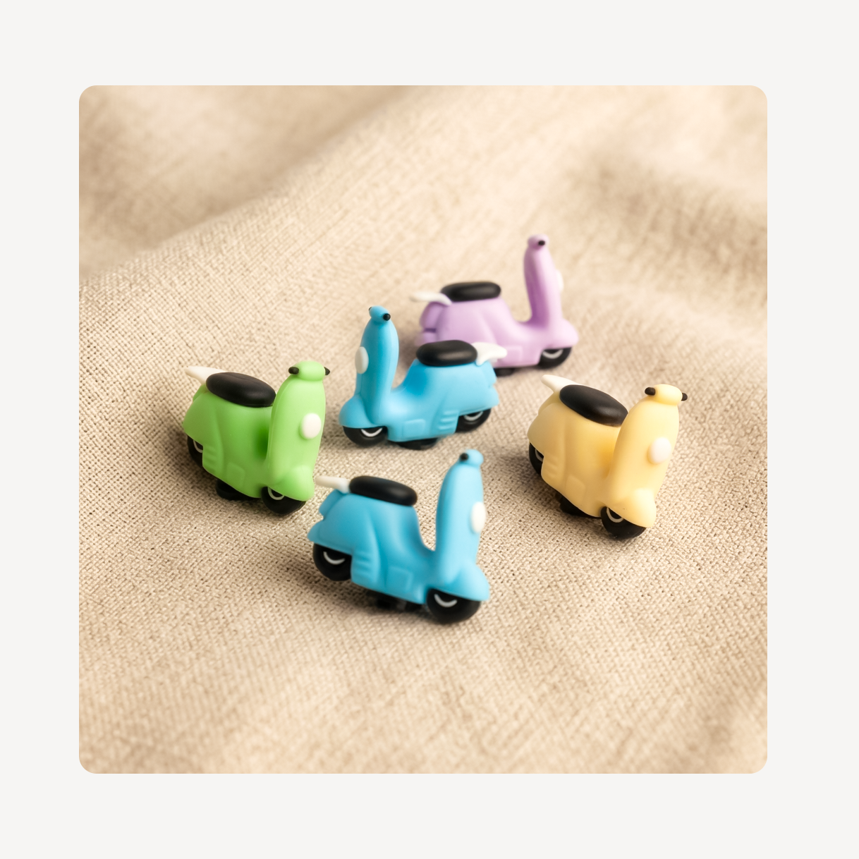 Five small scooter-shaped clog charms in green, light blue, lavender, and pale yellow, arranged on a beige textured fabric background.