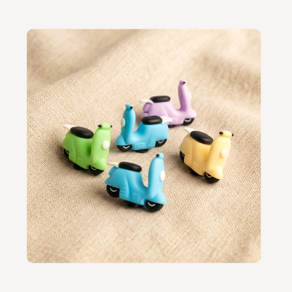 Five small scooter-shaped clog charms in green, light blue, lavender, and pale yellow, arranged on a beige textured fabric background.