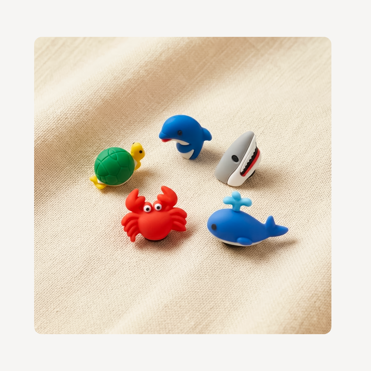 Five small sea-themed clog charms arranged on a beige fabric background: a green turtle with a yellow head, a blue dolphin, a gray shark with red and white teeth, a red crab, and a blue whale with a light blue fin.