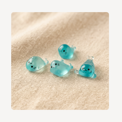 Four small translucent aqua seal-shaped clog charms with embroidered black facial features, arranged on a beige textured fabric background.