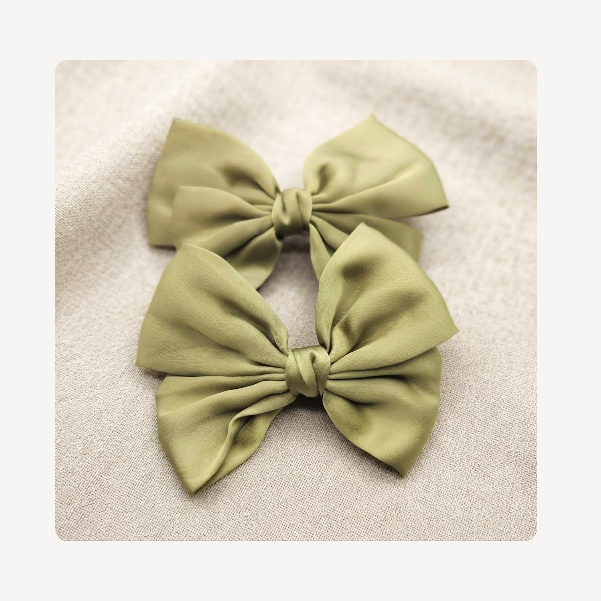 Two olive green satin sneaker bows arranged on a textured beige fabric background.